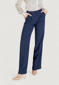 Pantalons taillés navy à taille haute, avec des plis devant et des poches latérales, associés à une chemise légère et transparente à boutons et des escarpins nude.