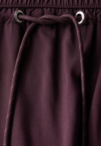 Tasche in bordeaux con cintura elasticizzata e lacci. Presenta una superficie texturizzata e occhielli metallici con cordino scuro. Finitura in tessuto liscia.
