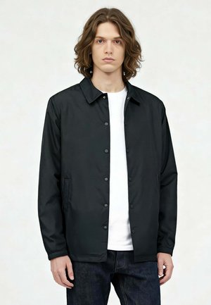 Jonge man met schouderlang bruin haar draagt een zwarte jas met knopen over een wit shirt en donkere spijkerbroek, staande tegen een lichte achtergrond.