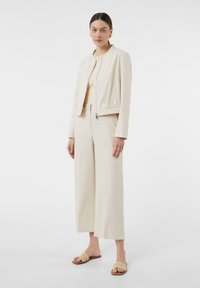 Kremowa, strukturalna kurtka z okrągłym dekoltem i zamkiem, zestawiona z szerokimi kremowymi spodniami i plecionymi klapkami.