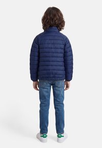 Παιδί με καστανά σγουρά μαλλιά που φοράει σκούρο μπλε μπουφάν τύπου puffer, μπλε τζιν και λευκά αθλητικά με πράσινες λεπτομέρειες, στέκεται γυρισμένο με την πλάτη.