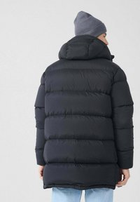 Persona con una chaqueta de invierno negra acolchada con capucha y un gorro gris, vista desde atrás contra un fondo claro.