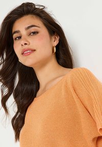 Suéter de punto naranja con textura acanalada y escote amplio. La modelo lleva puestos pendientes de aro dorados. El cabello cae en ondas sueltas.