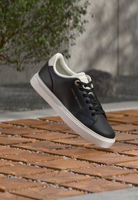 Baskets basses noires et blanches avec semelle blanche, lacets noirs et logo doré "Tommy Hilfiger", flottant au-dessus d'une surface en bois à motifs.