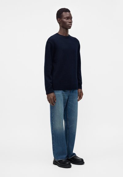 Homme debout portant un pull bleu marine foncé, un jean bleu droit et des chaussures noires épaisses sur un fond clair uni.