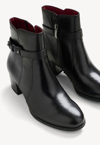 Melnas ādas potītes zābaki ar gludu apdari, norādītu pirkstu daļu, zemu blokveida papēdi, sānu rāvējslēdzējiem un dekoratīvu siksniņu ar kniedēm.