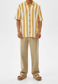 Kortärmad skjorta med gula och vita zigzag-ränder, knappstängning, tillsammans med ljusbeiga byxor och beige sandaler.