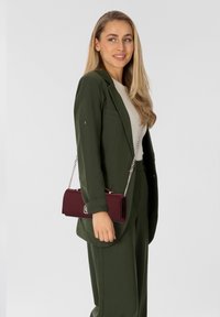 Blazer verde y pantalones a juego con una blusa blanca, sosteniendo un bolso cilíndrico burdeos con una correa de cadena, que presenta herrajes plateados.