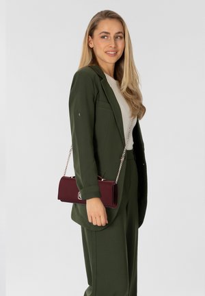 Groene blazer en bijpassende broek met een wit topje, een bordeauxrode cilindrische tas met een kettingriem vasthoudend, met zilveren accessoires.