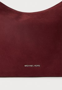 Bordo kožni kovčeg s glatkom teksturom, zaobljenim gornjim rubom i srebrnim logotipom "MICHAEL KORS" na dnu.