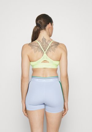 Bustier sportiv de culoare galben-neon, cu bretele încrucișate și detaliu tăiat pe spate, asortat cu pantaloni scurți albaștri deschiși cu accente verzi și logo.