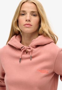 Felpa con cappuccio rosa, dotata di cordino, con una finitura texture e logo ricamato sul petto. Tessuto liscio e vestibilità rilassata.