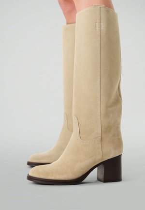 Beige Wildlederstiefel bis zu den Knien mit quadratischer Zehenpartie, sichtbaren Nähten und einem klobigen Holzblockabsatz. Glatte Textur mit minimalen Akzenten.