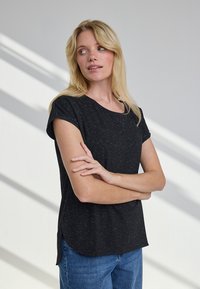 Blond naine käed rinnal, seljas must lühikeste varrukatega särg ja sinised teksad, vaatab oma vasakule heleda tausta ees.