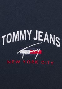 Marinfärgad tyg har vit broderad text "TOMMY JEANS" och "NEW YORK CITY," med en röd, vit och blå randig accent nedanför.