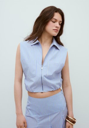 Žena oblečená v modro-bielom pruhovanom crop tope bez rukávov so zipsom a zodpovedajúcej sukni s vysokým pásom, doplnenej náramkami.