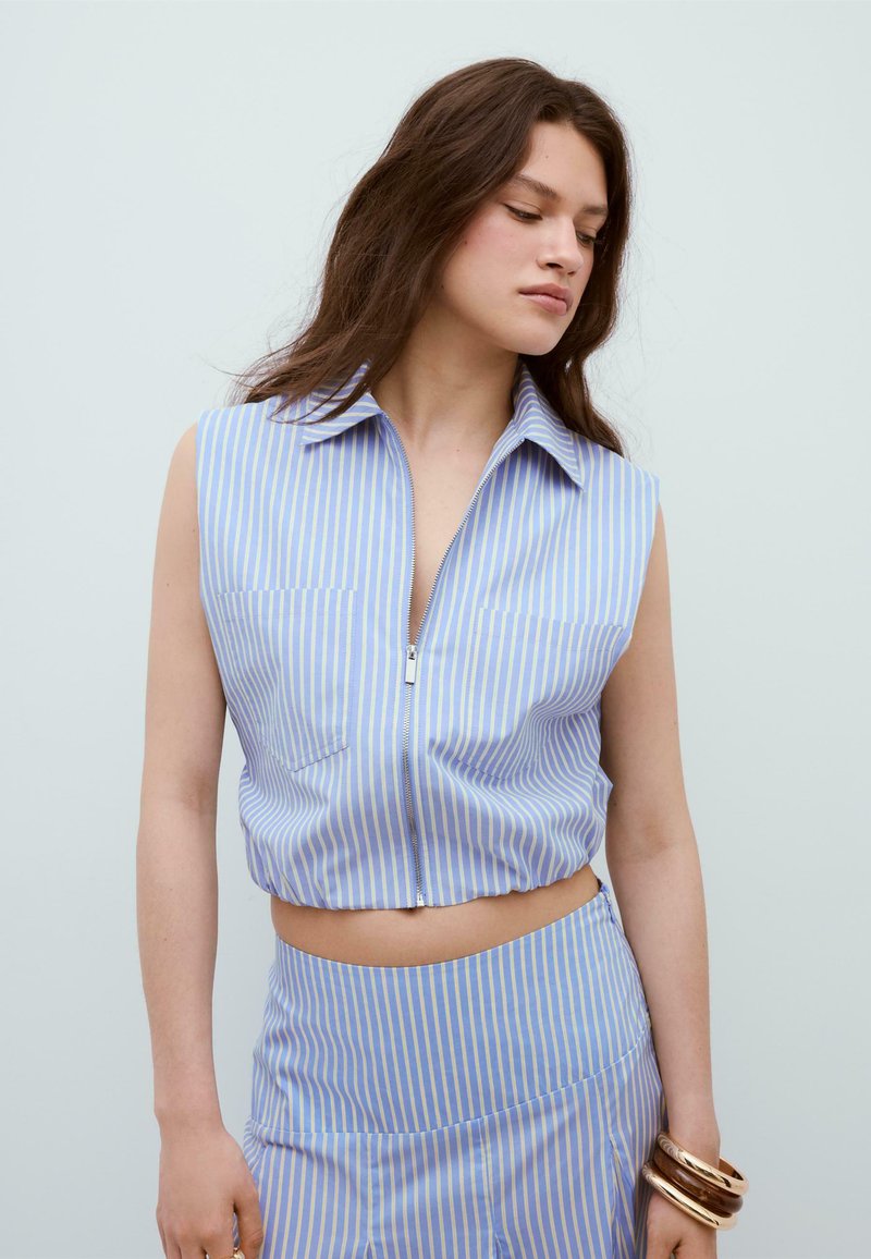 Femme portant un crop top sans manches rayé bleu et blanc avec une fermeture éclair et une jupe taille haute assortie, accessoirisée de bracelets.