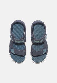 Blå textilsandal med justerbara remmar, texturerad fotbädd, vita sulor och Timberland-logotyp i ljusblått.