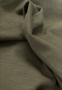 Zbliżenie na miękką, oliwkowozieloną tkaninę o delikatnej fakturze dzianiny oraz naturalnych fałdach tworzących głębokość i cień.