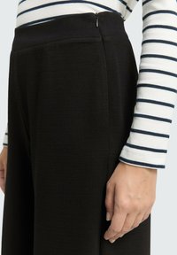 Pantalons noirs coupés avec un tissu texturé, dotés d'une fermeture éclair sur le côté. Un haut rayé à manches longues est partiellement visible au-dessus.