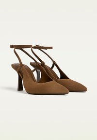 Décolleté in suede marrone, con punta a punta e cinturini sottili alla caviglia, dotate di tacco a blocco di altezza media, su sfondo bianco.