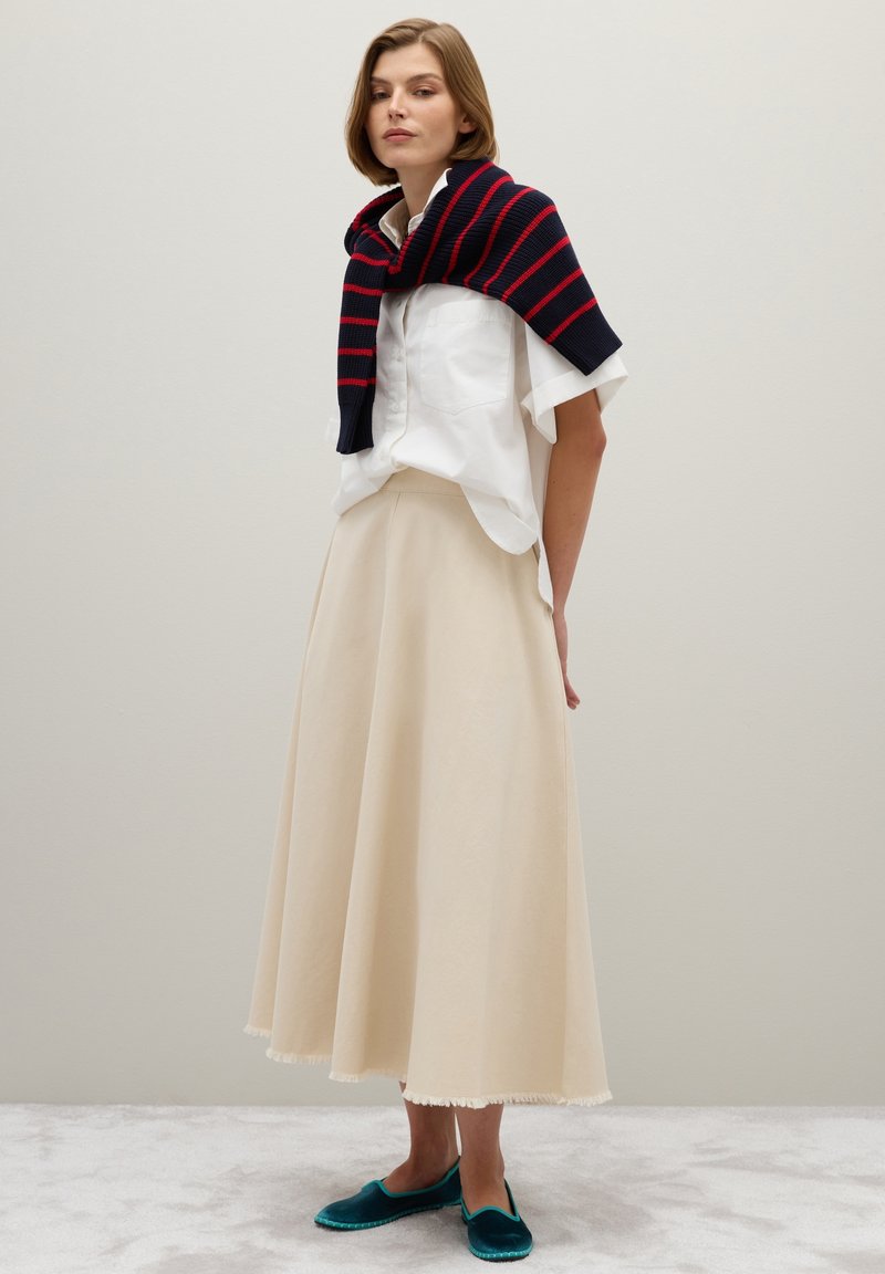 Camisa blanca de botones, falda beige fluida con dobladillo con flecos, suéter de rayas azul marino caído sobre los hombros, zapatos planos color verde azulado.