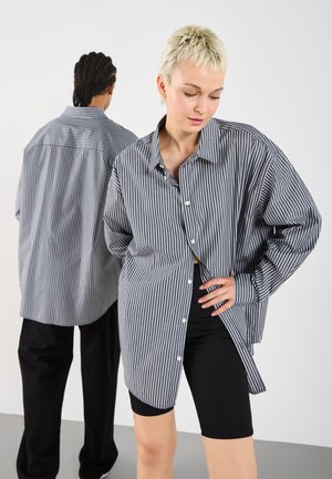 Stripet skjorte med knapper i svart og hvit, har oversized passform, lange ermer og buet hem. Svart shorts brukes under.
