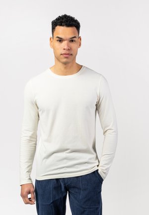 Langärmliges, cremefarbenes T-Shirt aus einem weichen, leichten Stoff, mit einem runden Ausschnitt und einem lockeren Schnitt. Kombiniert mit dunkelblauen Hosen.
