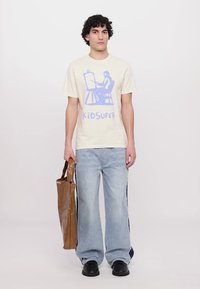 Beige T-Shirt mit einem hellblauen Grafikdesign und Text, kombiniert mit hellblauen Jeans. Das Model hält eine braune, strukturierte Tragetasche. Schwarze Schuhe.