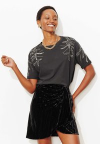 T-shirt noir avec broderie florale métallique sur les épaules, associée à une jupe en velours noir à rayures verticales et à une coupe portefeuille.