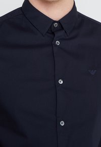 Mörkblå skjorta med spridd krage, som har en subtil textur, framknappar och en liten broderad logotyp på bröstet.