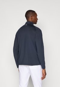 Pullover azul-marinho de manga longa em tecido liso, com colarinho alto, costuras nos ombros e um pequeno logotipo nas costas. Sem padrões visíveis.