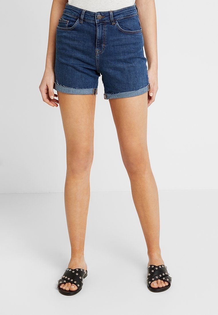 Tmavě modré džínové kraťasy s třepeným lemem a ohrnutými manžetami, doplněné černými nazouváky s cvočky.