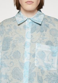 Camisa ligera y con estampado en tejido de azul claro y beige, presenta un diseño con cuello, botones frontales y un bolsillo en el pecho con una textura sutil.