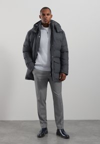 Grå vadderad jacka med huva, framfickor och dragkedja. Bärs över en ljusgrå tröja och grå skräddarsydda byxor. Svarta ankelboots.