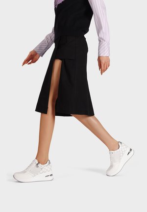 Sneaker bianche sportive con un motivo testurizzato, dettagli argentati e una suola ammortizzata, indossate con una gonna nera e un top a strati.
