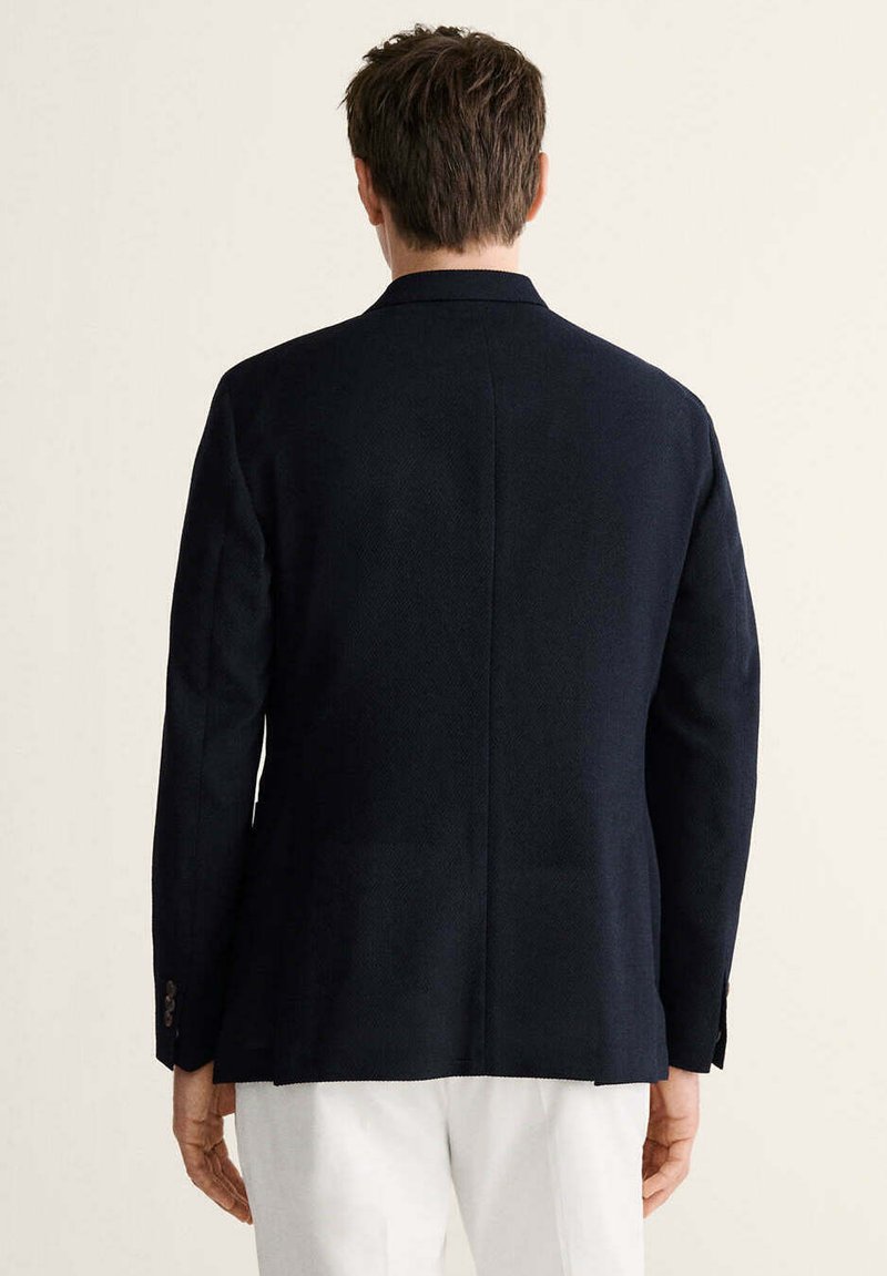 Hombre con blazer azul marino oscuro y pantalones blancos, visto desde atrás sobre un fondo claro y liso.