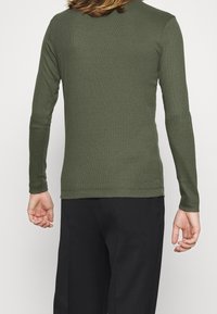 Langærmet, ribbet skjorte i olivengrøn, med rund halsudskæring og tætsiddende design, båret med sorte bukser.