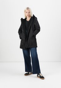 Black parka with a hood, featuring a fuzzy lining, front pockets, and a full-length zipper. Worn with wide-leg jeans and black sneakers.