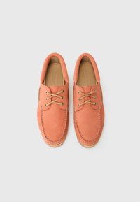 Zapatos de barco de ante rosa con cordones beige en contraste, punta redondeada y detalles de costura. La suela tiene un patrón de agarre texturizado.