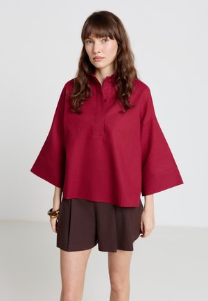 Frau mit langen braunen Haaren, die ein lockeres rotes Oberteil mit weiten Ärmeln und dunkelbraune plissierte Shorts trägt, steht vor einem einfarbigen Hintergrund.