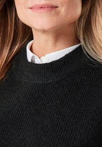 Schwarzer gerippter Pullover mit hohem Kragen, über einem weißen Hemd mit Kragen getragen. Fokus auf Textur und Stoffdetails.