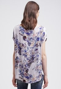 Kortärmad blus i lätt tyg med blommönster i nyanser av blått, lila och rosa, med rundad nederkant och avslappnad passform.