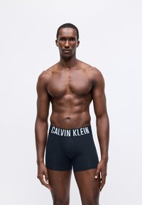 Fekete pamut boxer alsónadrág fehér "CALVIN KLEIN" logós derékszíjjal, rugalmas kialakítással. A modell mezítelen felsőtesttel látható, sima bőrrel.