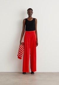 Rode wijde broek met plooien, gecombineerd met een zwarte tanktop. Het model houdt een rood-wit gestreept gebreid trui vast. Soepele stofstructuur.