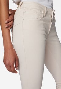 Jean slim beige en denim extensible. Comprend deux poches avant avec accents métalliques et détails de couture discrets.