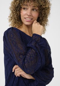 Námořnická modrá krajková blůza s diamantovými vzory, volný střih a nabírané rukávy. Obsahuje texturované detaily a jemný, průhledný vzhled.