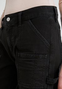 Pantalones cargo negros con una silueta ajustada, múltiples bolsillos, textura resistente y detalles de costura en contraste en los lados y la cintura.