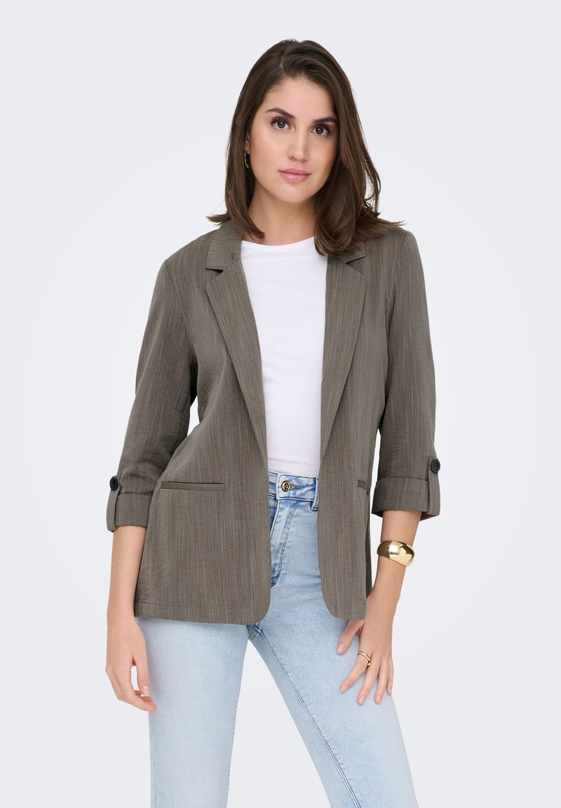Blazer léger vert olive en tissu légèrement texturé, avec des revers crantés, des manches retroussées et deux poches avant, porté avec un jean bleu clair.