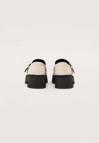 Beige Wildlederschuhe mit einer klobigen schwarzen Gummisohle, ausgestattet mit einem Rückriemen und einem leichten Absatz, konzipiert für einen sicheren Sitz und Langlebigkeit.